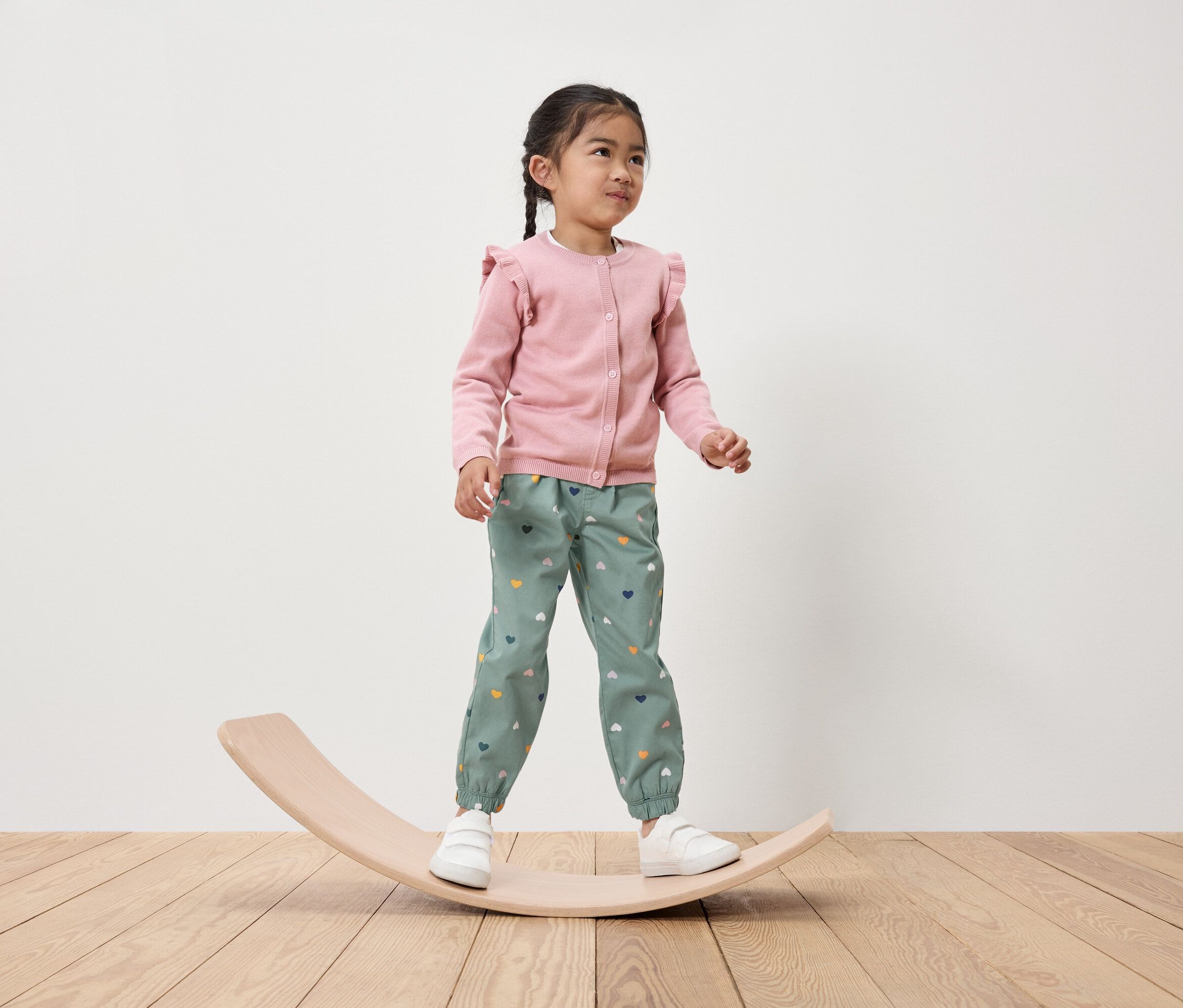 Holčička stojí na dřevěné balanční desce, má na sobě růžový kardigan a zelené kalhoty se srdíčky.