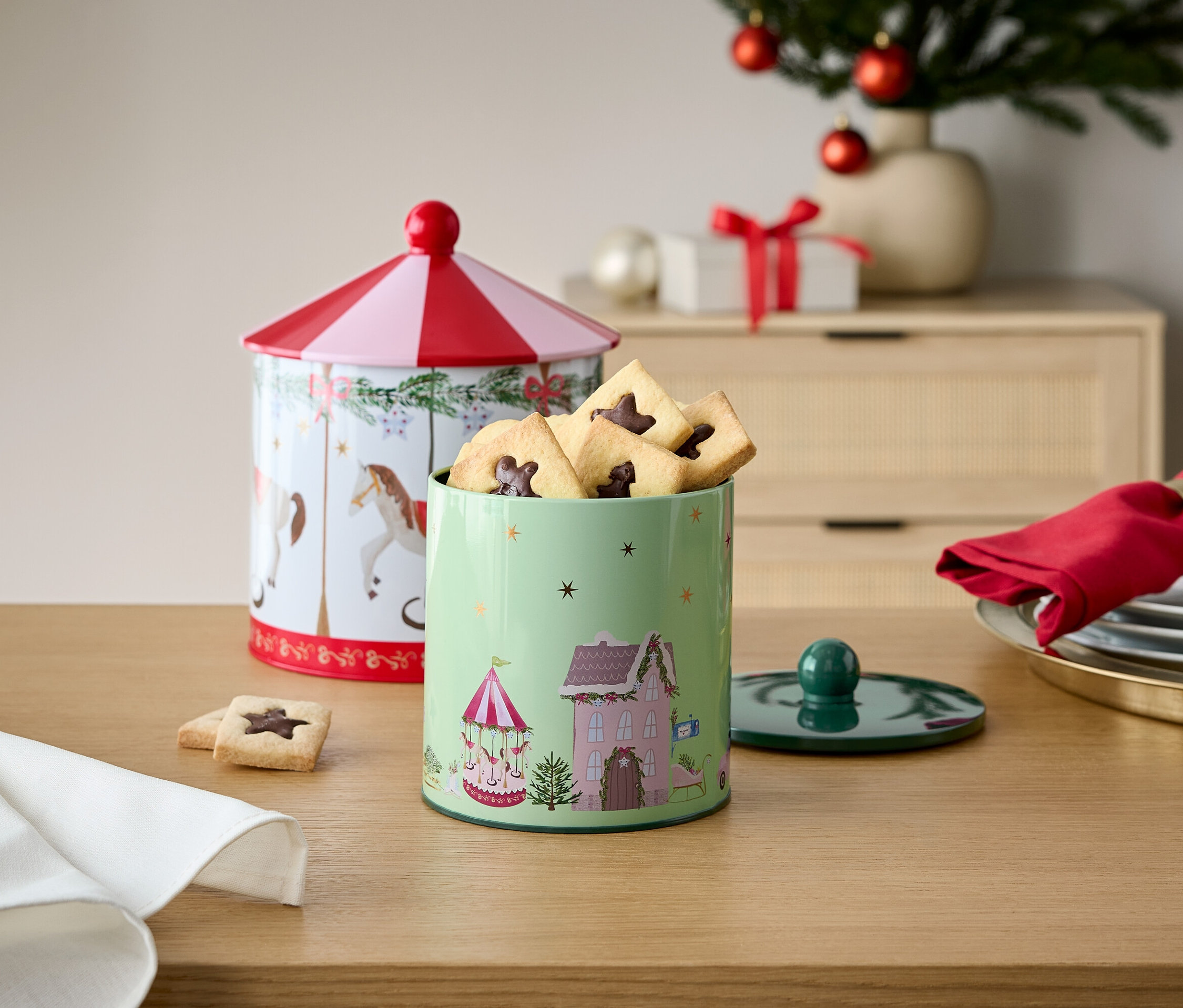 Dva zdobené plechové dózy na sušenky na dřevěném stole s vánoční výzdobou v pozadí.