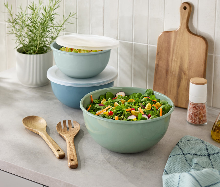 Na lince stojí mísa se salátem, dřevěný příbor, slánka, láhev oleje, prkénko a rostlina v květináči.