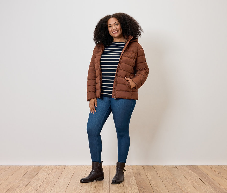 Žena s kudrnatými vlasy pózuje v hnědé prošívané bundě, modrých džínách a hnědých botách.