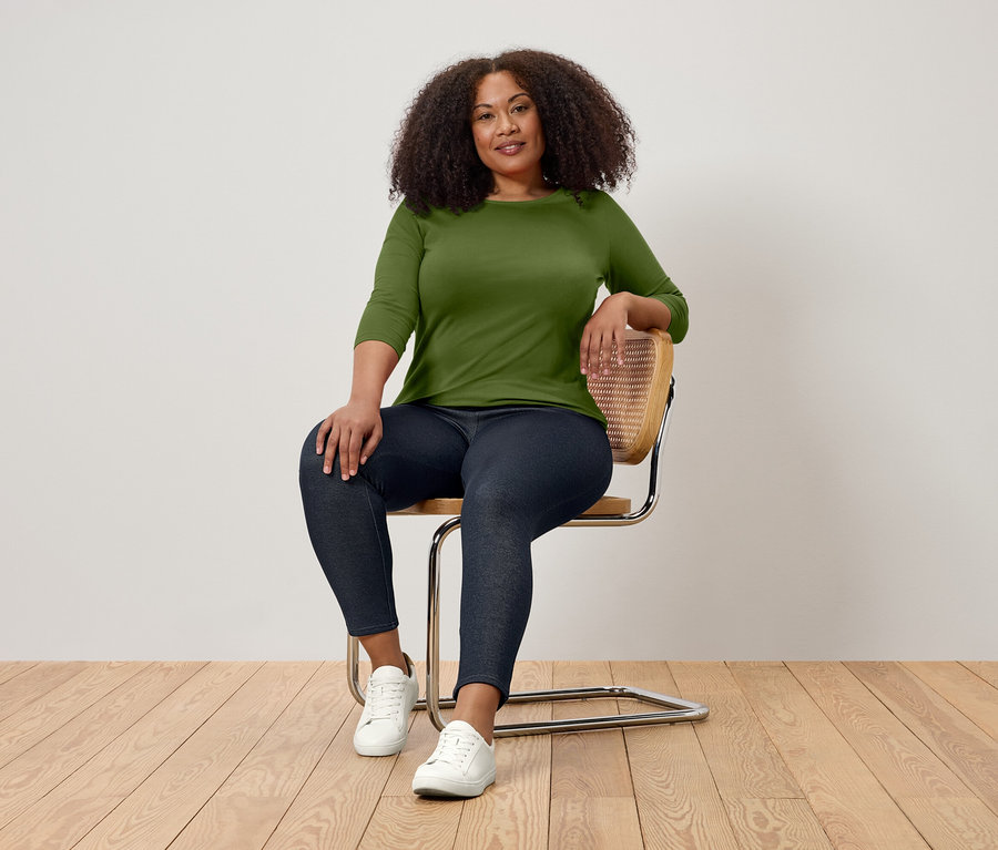 Žena s kudrnatými vlasy sedí na židli, má na sobě zelené tričko s tříčtvrtečním rukávem, tmavé džíny a bílé tenisky.