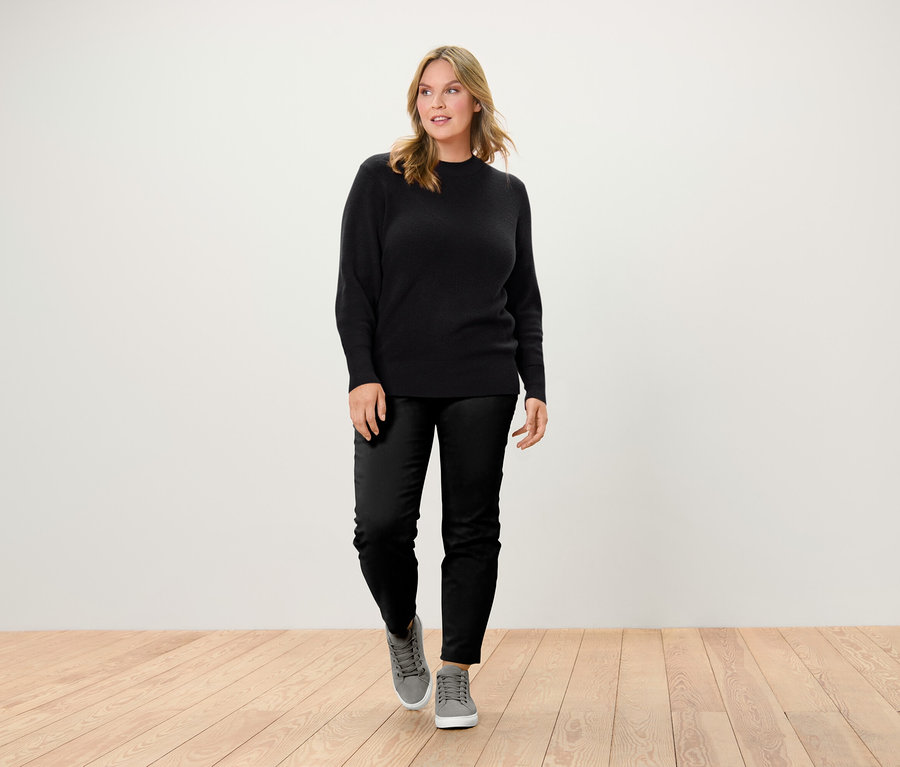 Žena kráčející po dřevěné podlaze má na sobě černý svetr, černé kalhoty a šedé tenisky.