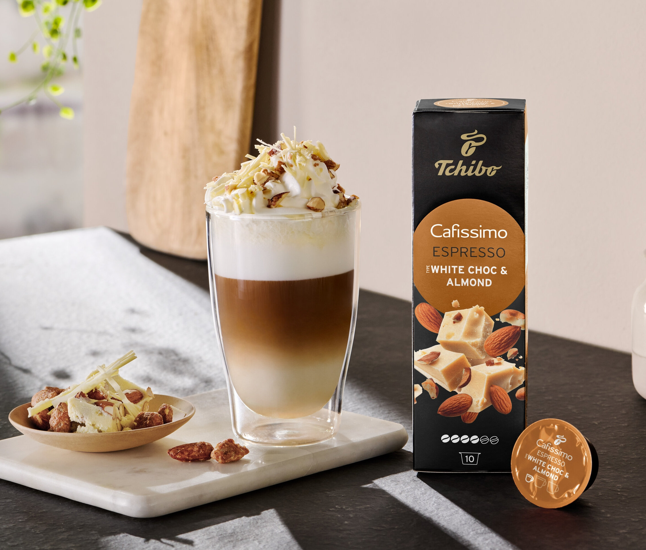 Vrstvené latte s šlehačkou a posypem vedle balení Cafissimo Flavoured Espresso - White Choc & Almond.
