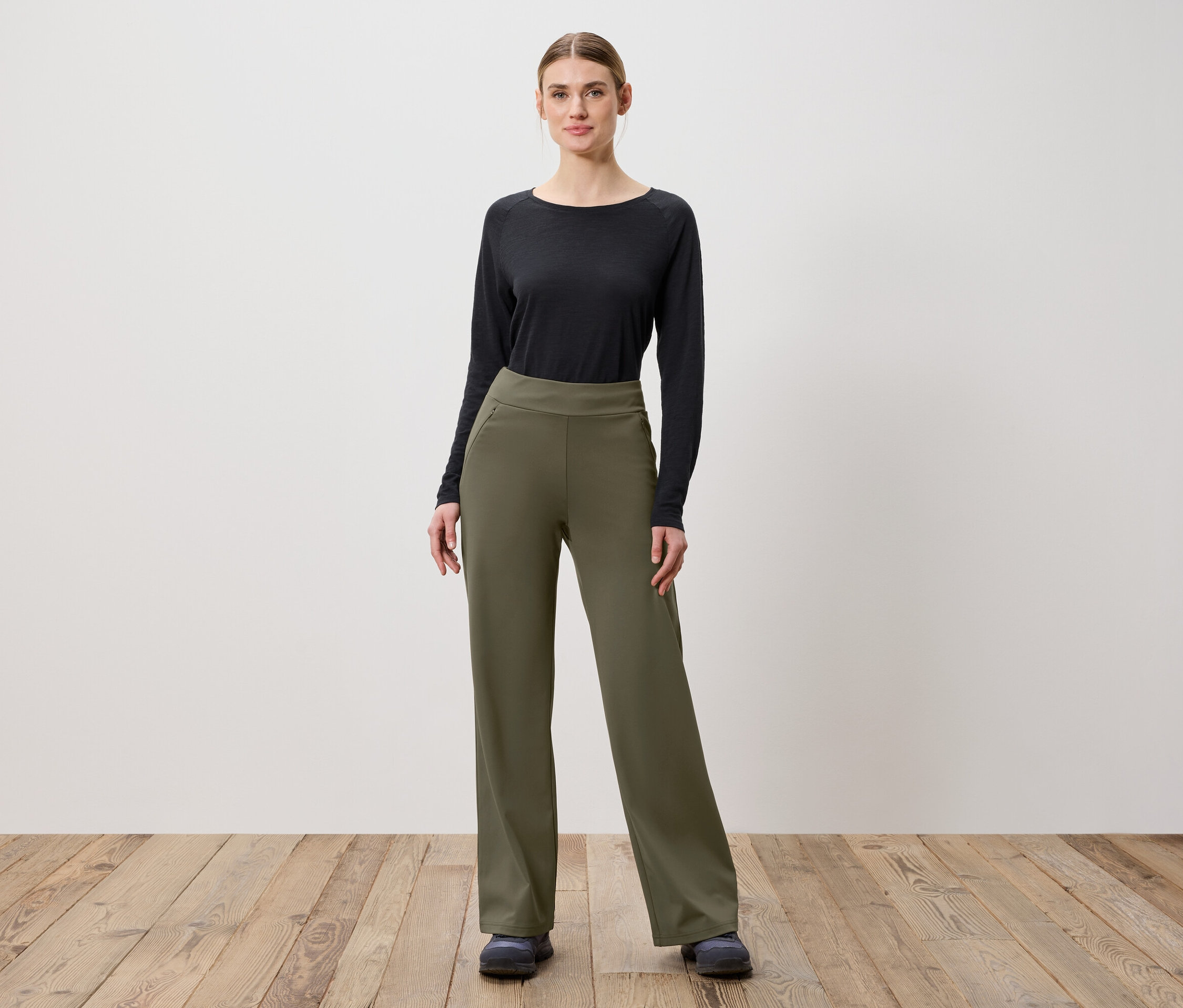 Žena pózuje na dřevěné podlaze v olivově zelených kalhotách a černém topu s dlouhým rukávem.