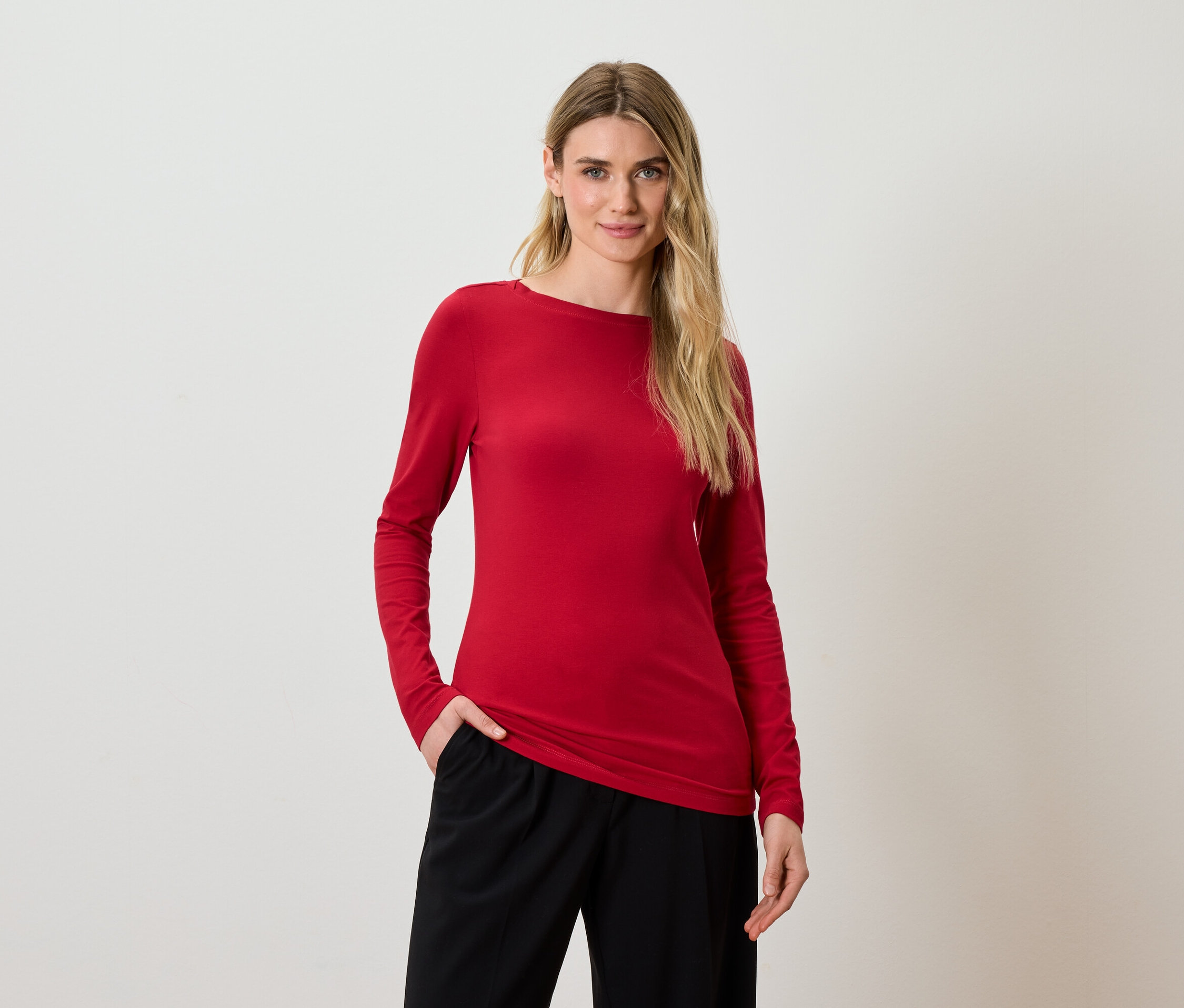 Žena s blond vlasy pózuje v červeném tričku s dlouhým rukávem a černých kalhotách.