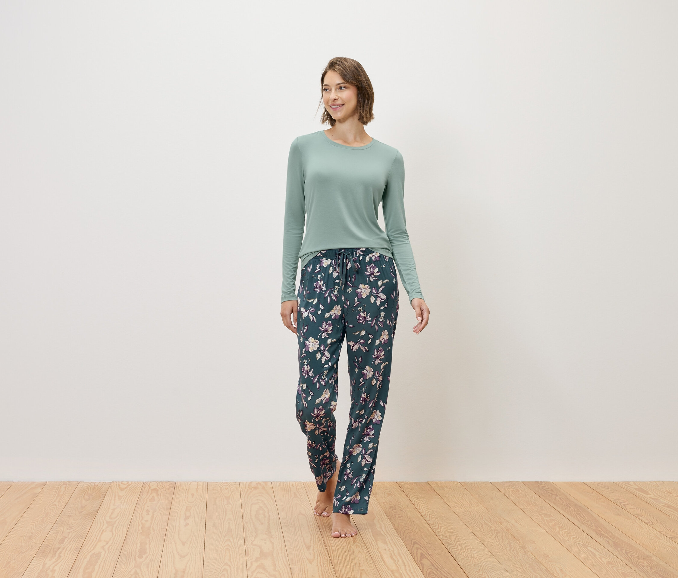 Žena jde bosá a má na sobě mátový svetr a tmavě modré pyžamové kalhoty s květinovým vzorem.