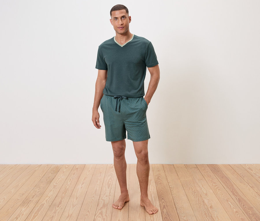 Muž stojí bosý na dřevěné podlaze a má na sobě zelené pyžamo s krátkým rukávem a šortky.