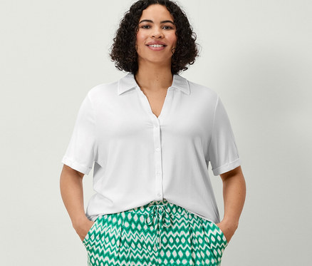 Žena stojí a má ruce v kapsách 3/4 žerzejových kalhot s potiskem a bílého halenkového trička s knoflíkovou légou.