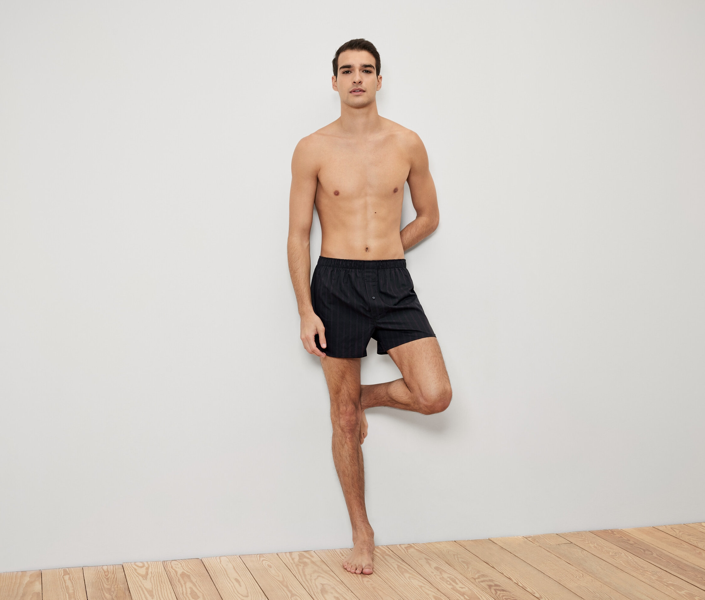 Muž pózuje opřený o zeď v černých pruhovaných boxerkách.
