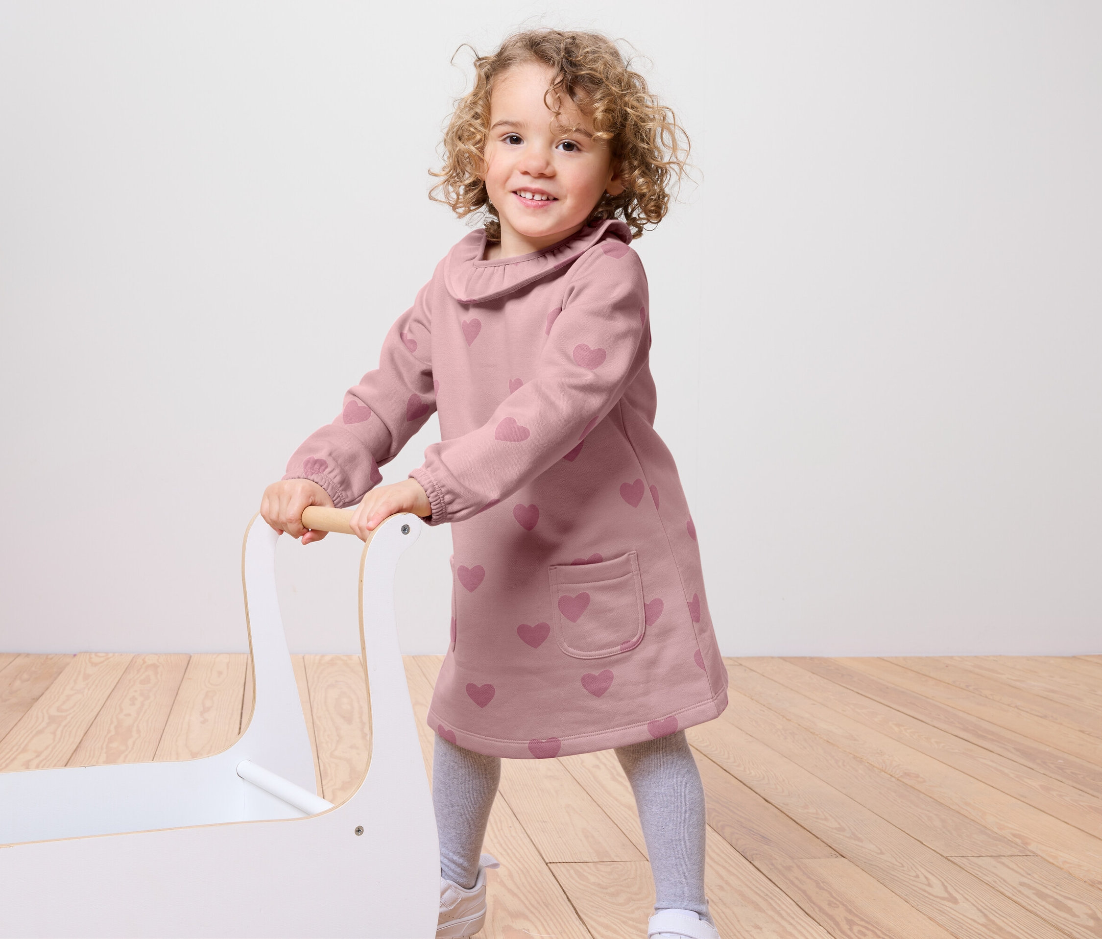 Usmívající se holčička tlačí bílý vozík na hračky. Má na sobě růžové šaty se srdíčky, šedé legíny a bílé tenisky.
