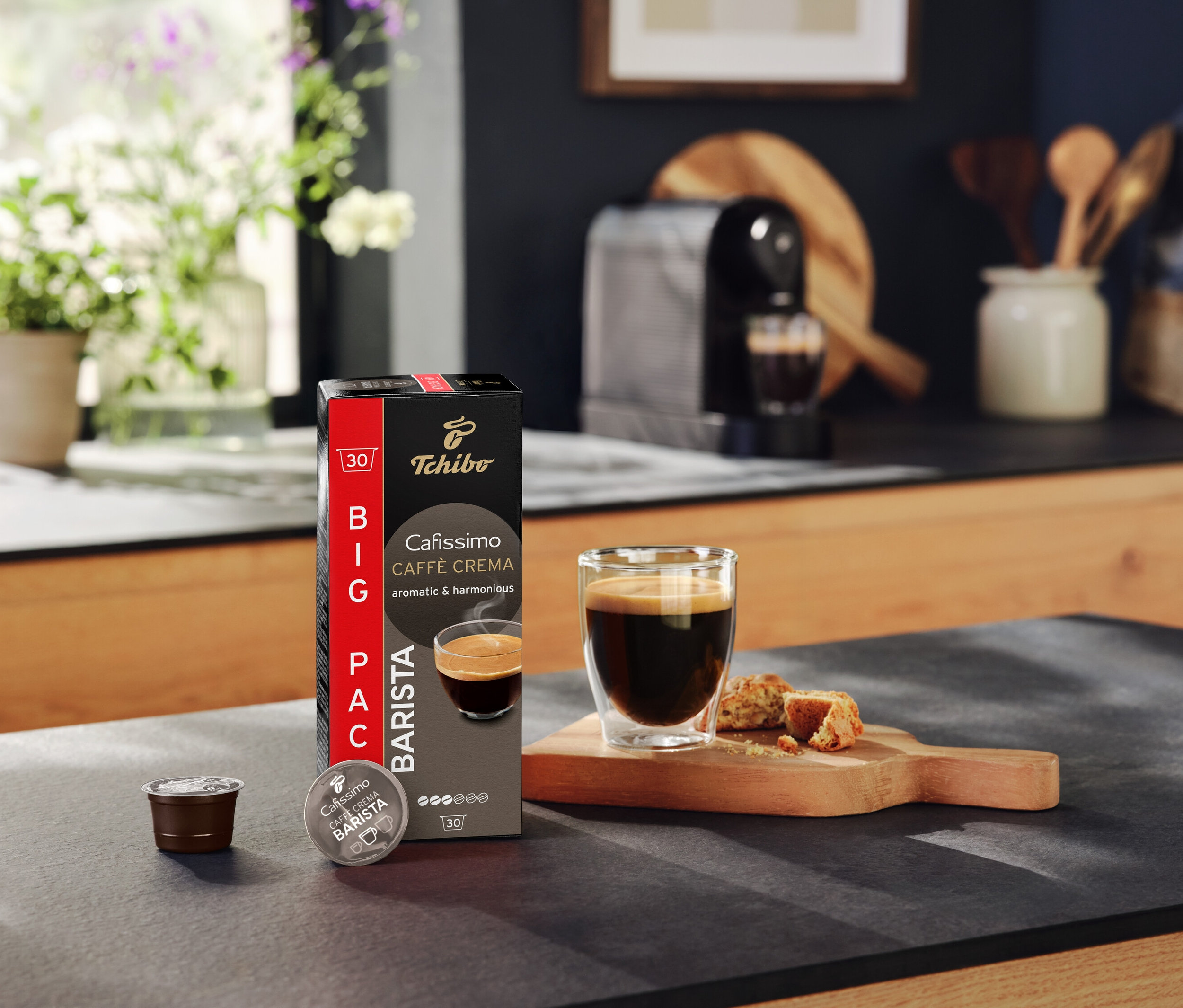 Na kuchyňské lince stojí balení Tchibo Cafissimo Caffè Crema, kapsle, šálek kávy a pečivo.