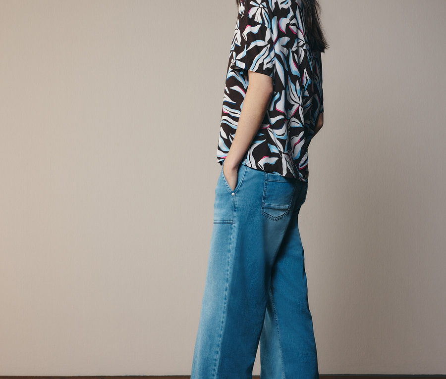 Žena v vzorované halence a rozšířených džínách, dívající se do strany.