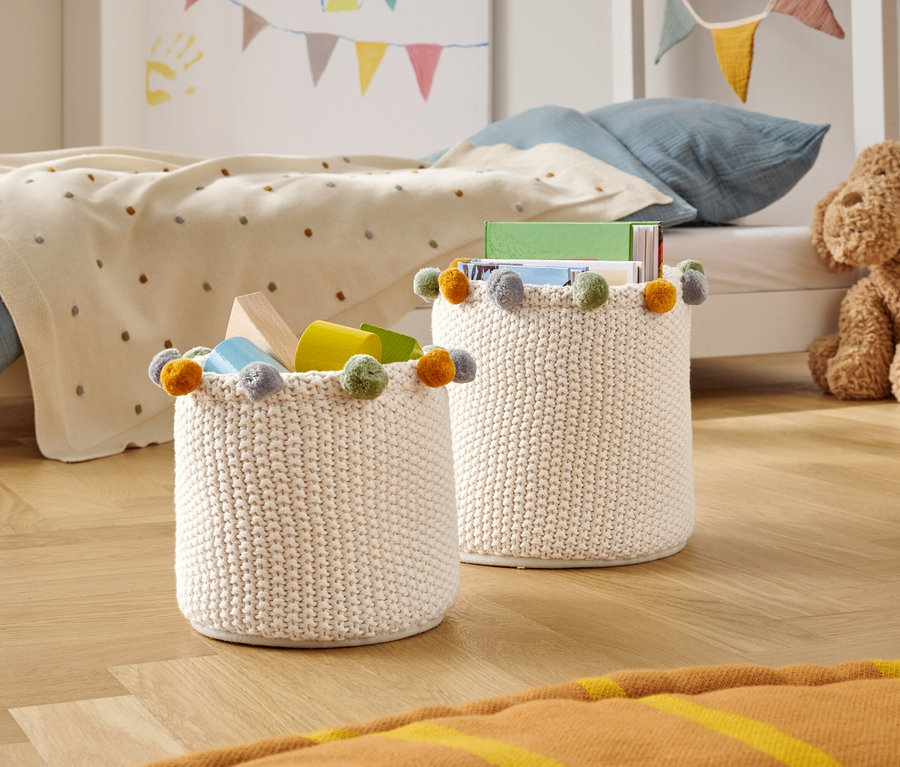 Dva pletené úložné koše s barevnými bambulkami stojí na dřevěné podlaze v dětském pokoji.