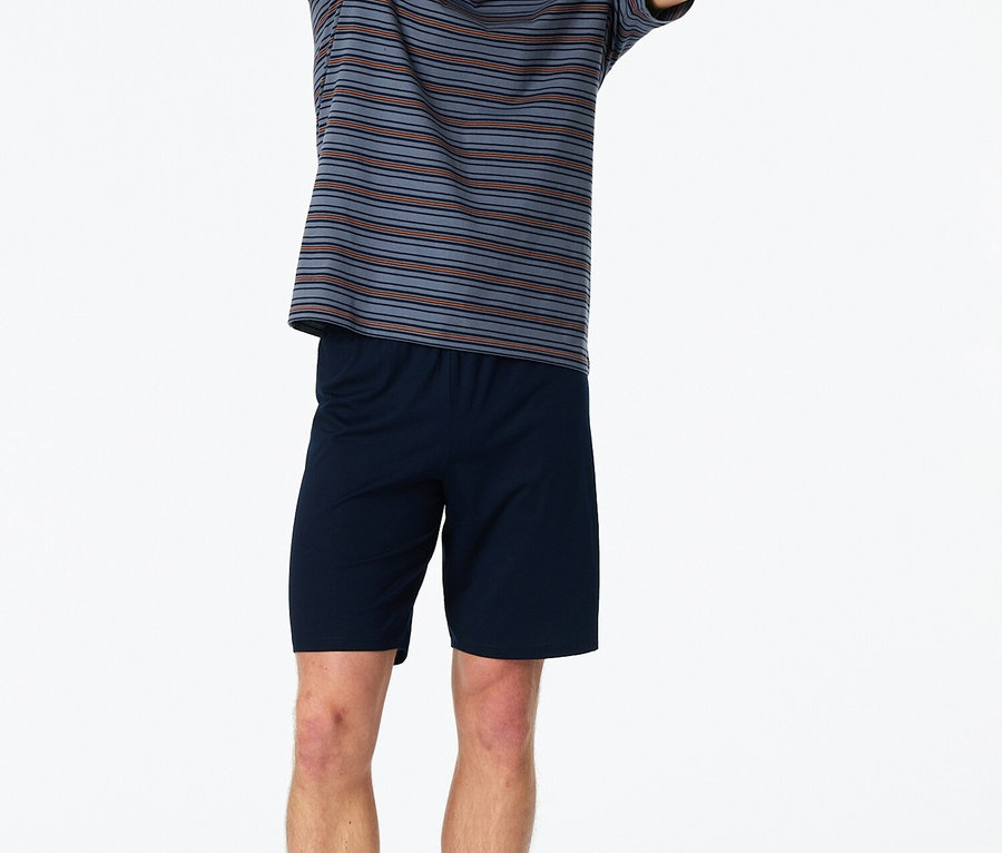 Muž stojí bosý se zvednutýma rukama v pruhovaném tričku a modrých šortkách na bílém pozadí.