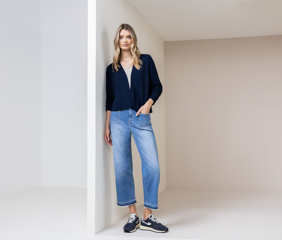 Žena se opírá o zeď, má na sobě tmavě modrý kardigan, modré džíny a tmavě modré boty.