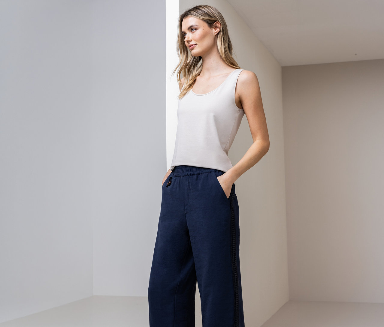 Žena s basic topem Street One a modrými kalhotami s rukama v kapsách před zdí.