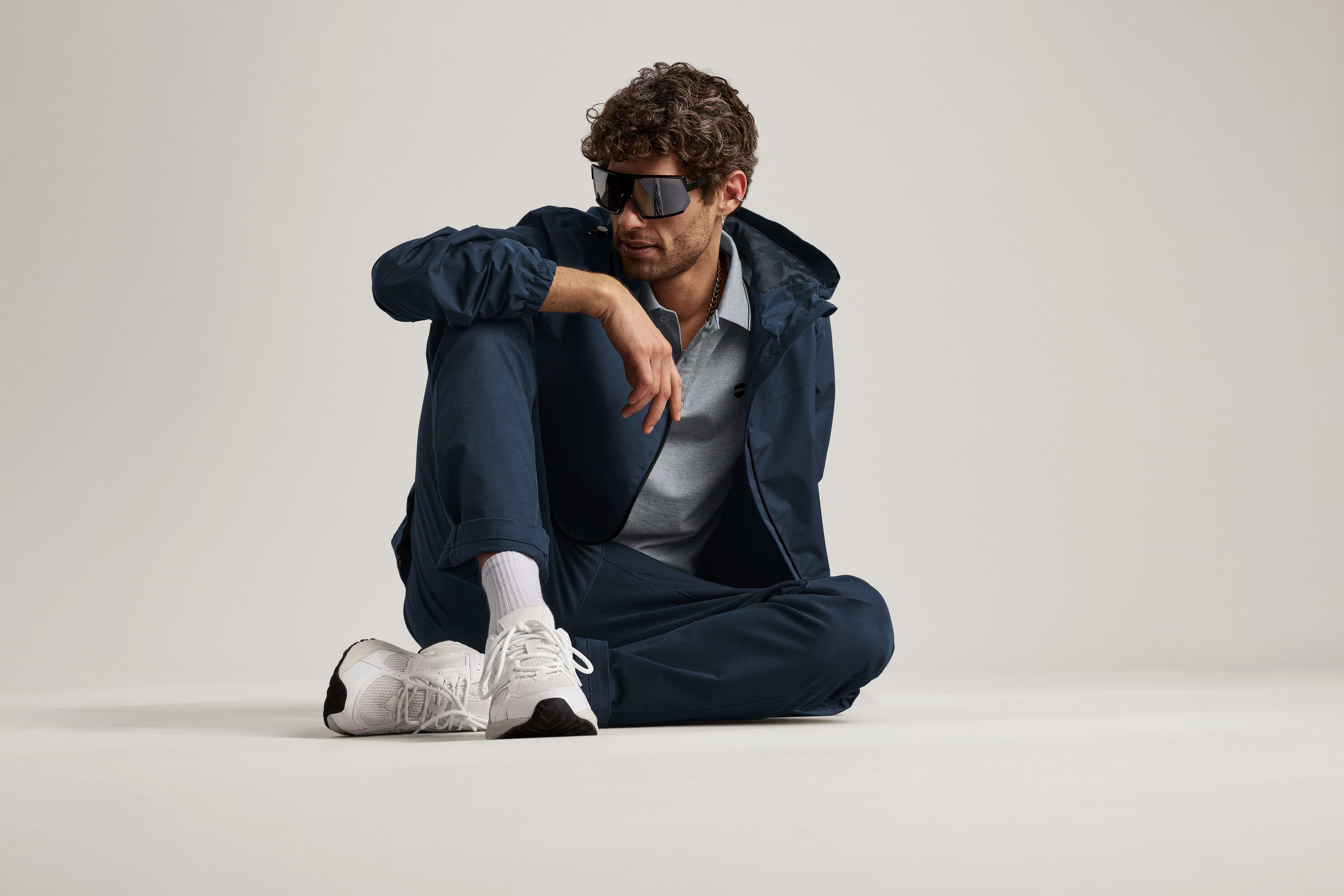 Muž sedí na zemi v námořnických chino kalhotách, bundě a slunečních brýlích.
