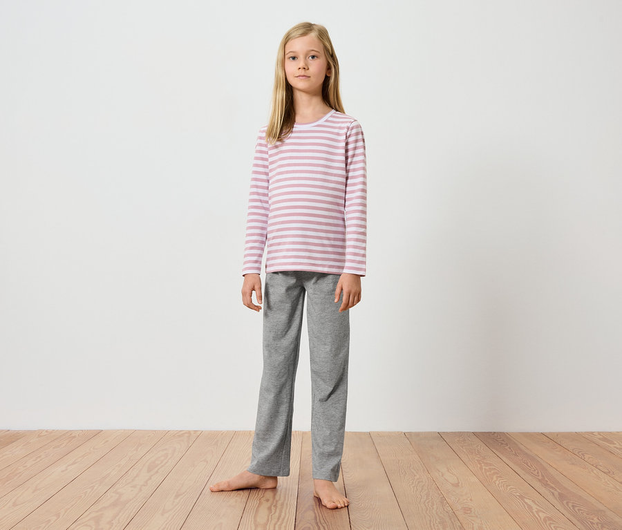 Mladá dívka stojí bosá na dřevěné podlaze před bílou zdí. Má na sobě růžovo-bílé pruhované tričko s dlouhým rukávem a šedé kalhoty.