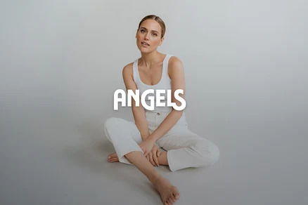 Žena sedí na podlaze, oblečená v bílém tílku a bílých kalhotách, se zkříženýma nohama, dívá se do kamery. Překryto textem "ANGELS".