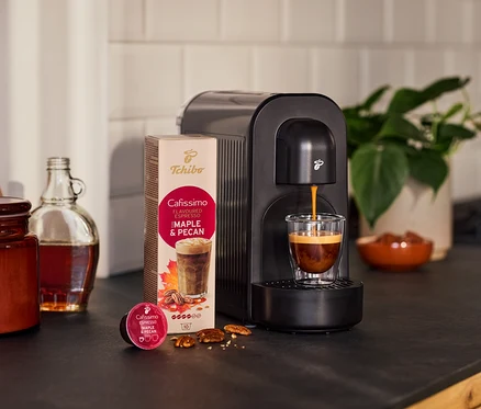 Černý kávovar vaří espresso do sklenice. Vedle balení kapslí s příchutí javoru a pekanových ořechů, volná kapsle, ořechy, láhev a rostlina.