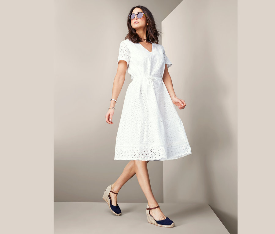 Žena kráčí a má na sobě bílé šaty s ažurovou výšivkou.