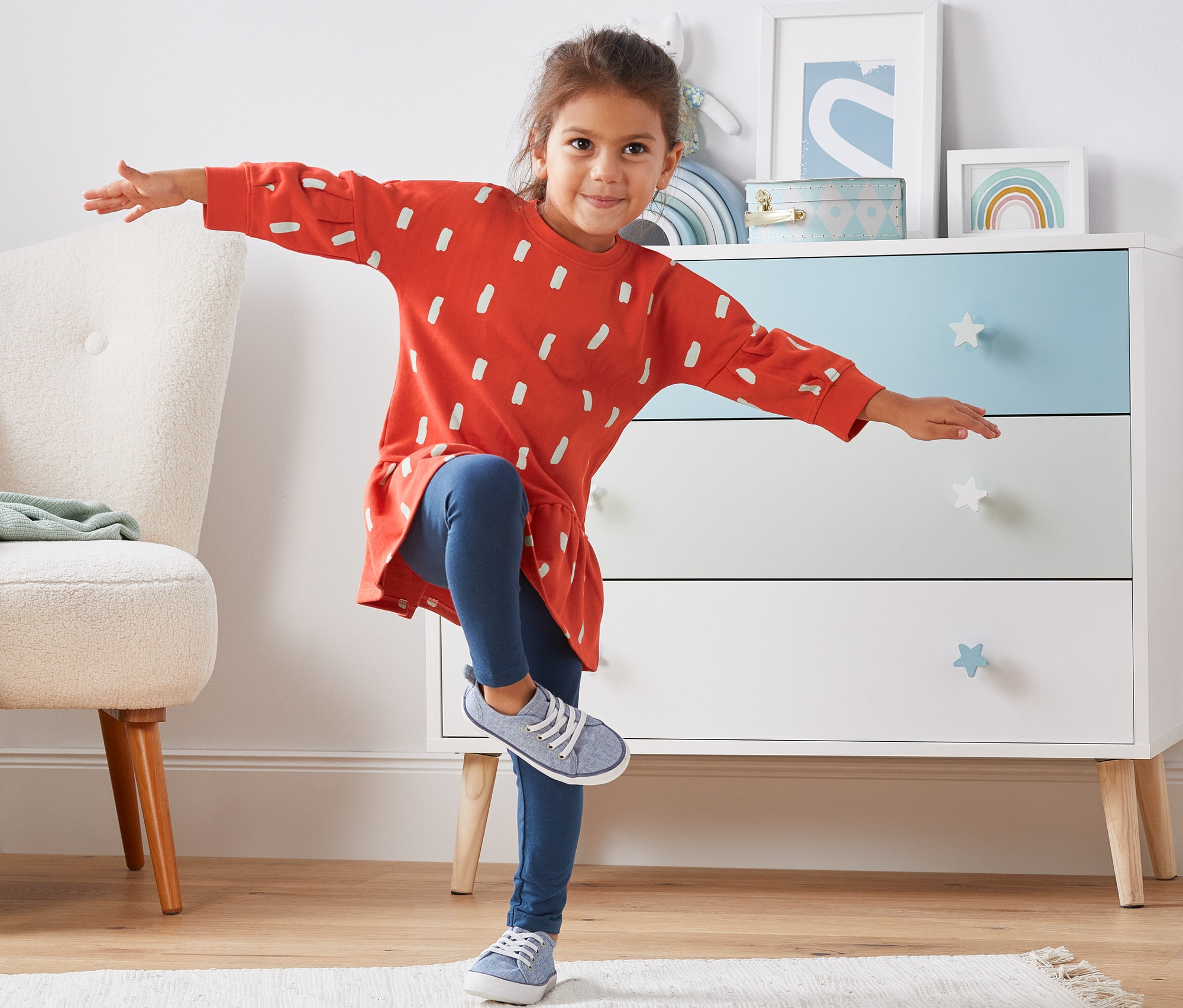 Dívka stojí na jedné noze a mává rukama. Má na sobě teplákové šaty z biobavlny.