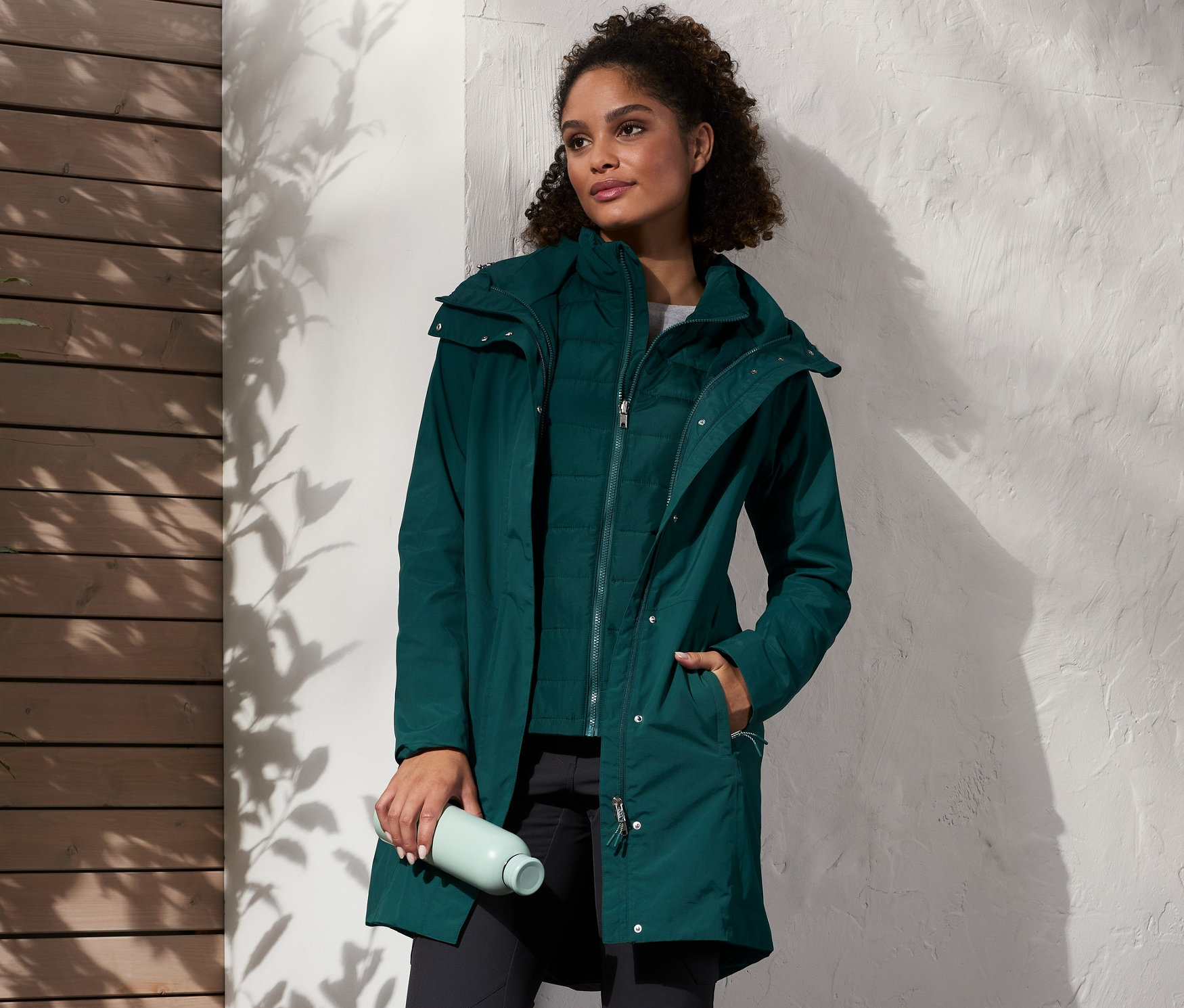 Žena stojí a má na sobě zelený kabát do deště s vestou, černé kalhoty a hnědé boty. V ruce drží láhev s vodou a vedle ní stojí batoh.