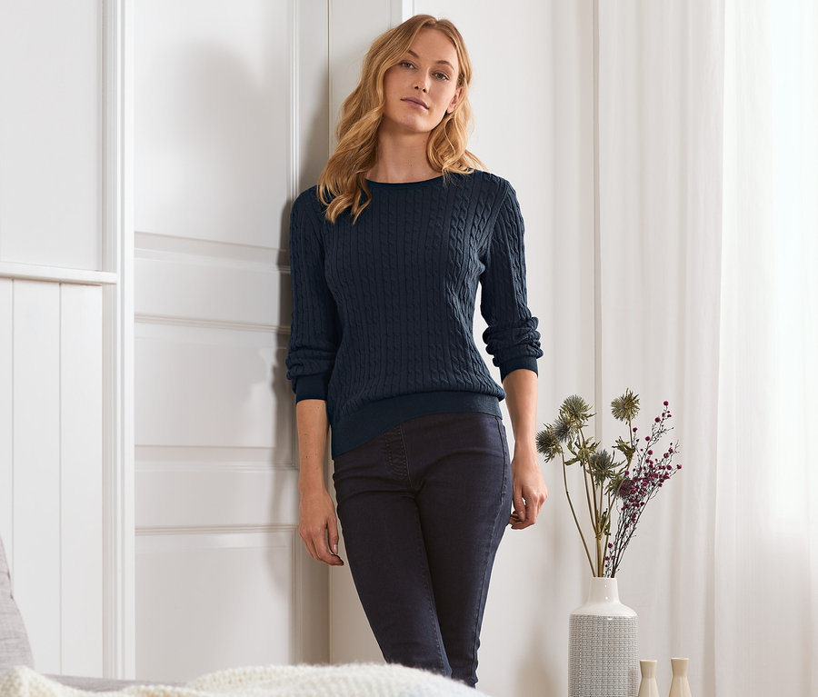 Žena opřená o dveře má na sobě modrý pletený svetr s copánkovým vzorem.