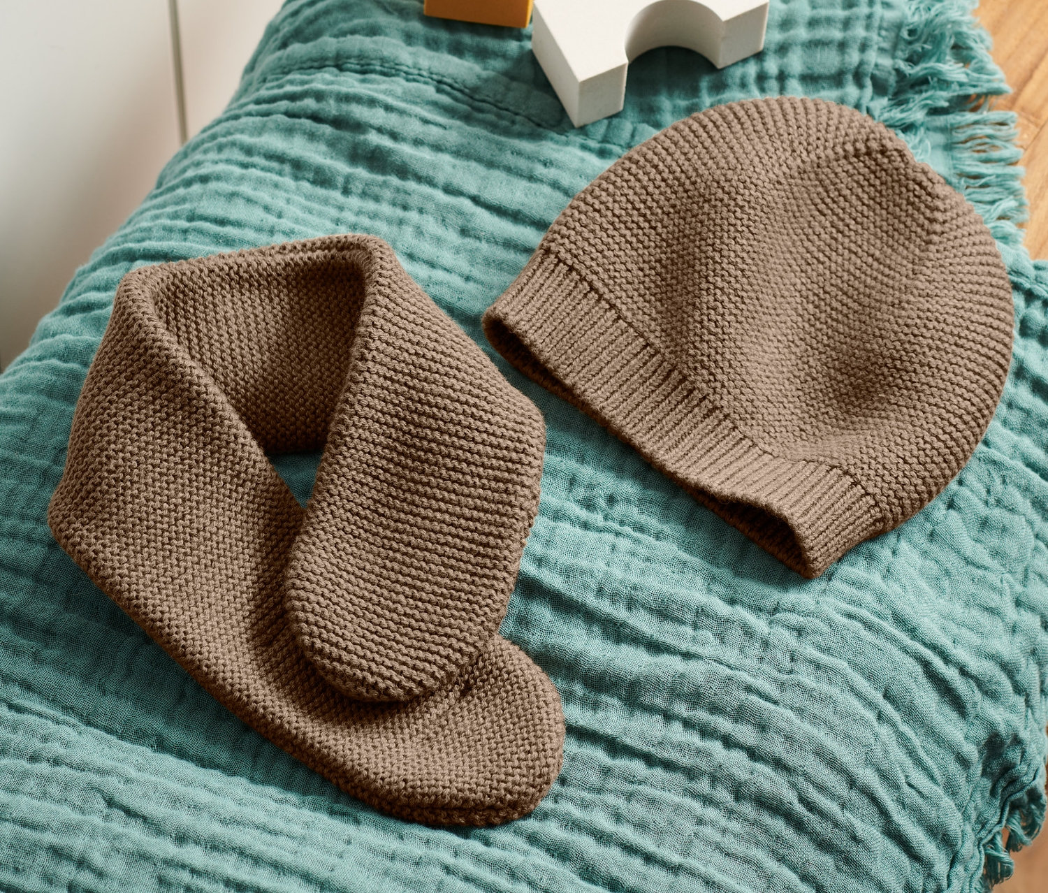 Hnědá pletená čepice a šála leží na dřevěné stoličce na modrozelené látce vedle bílých a žlutých kostek.