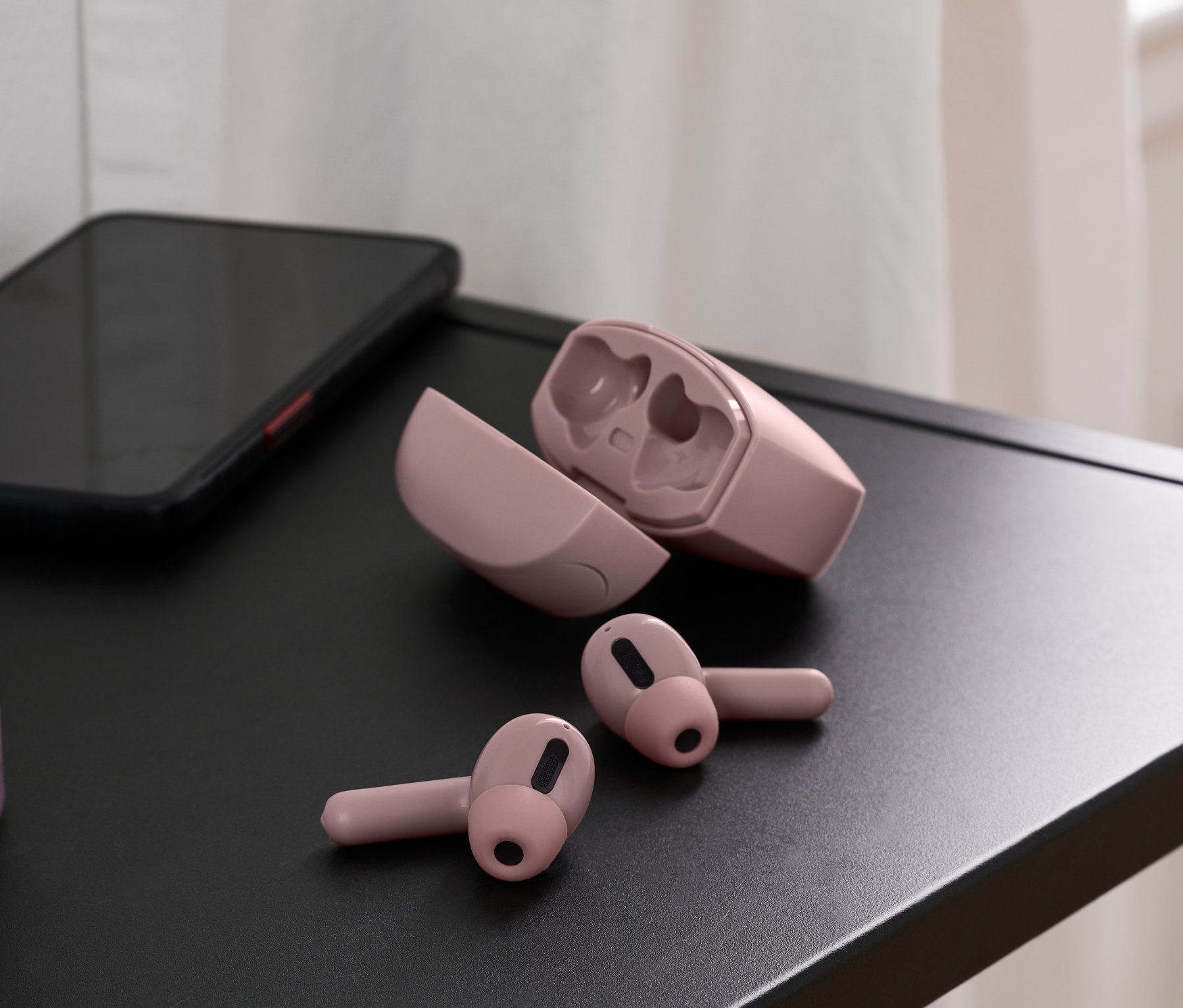 Na černé desce stolku leží světle růžová sluchátka do uší TWS, vedle nich je otevřené pouzdro a mobilní telefon. V pozadí váza.