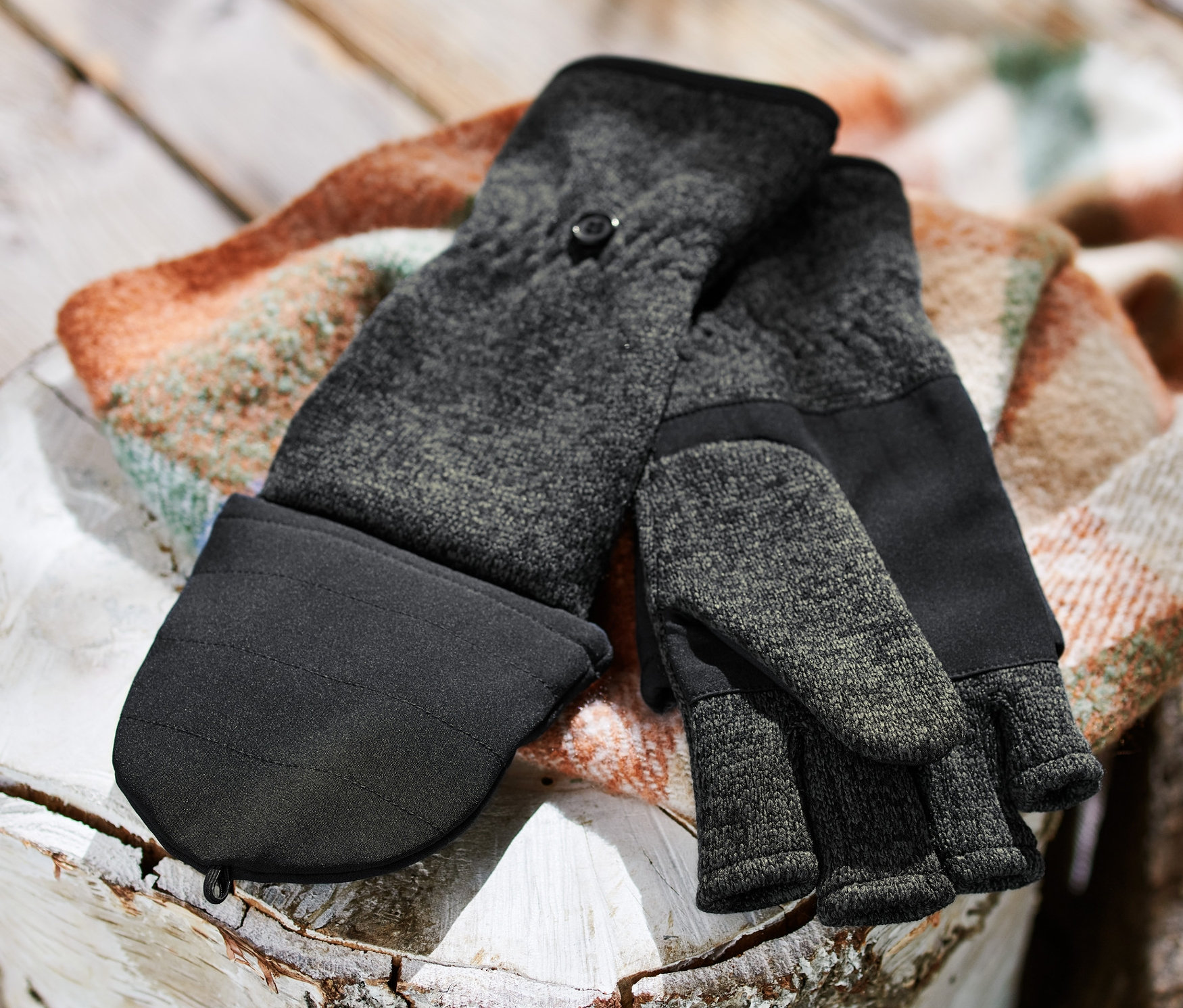 Pár odklápěcích palčáků z pleteného fleecu leží na dřevěném špalku vedle hromady dřeva.