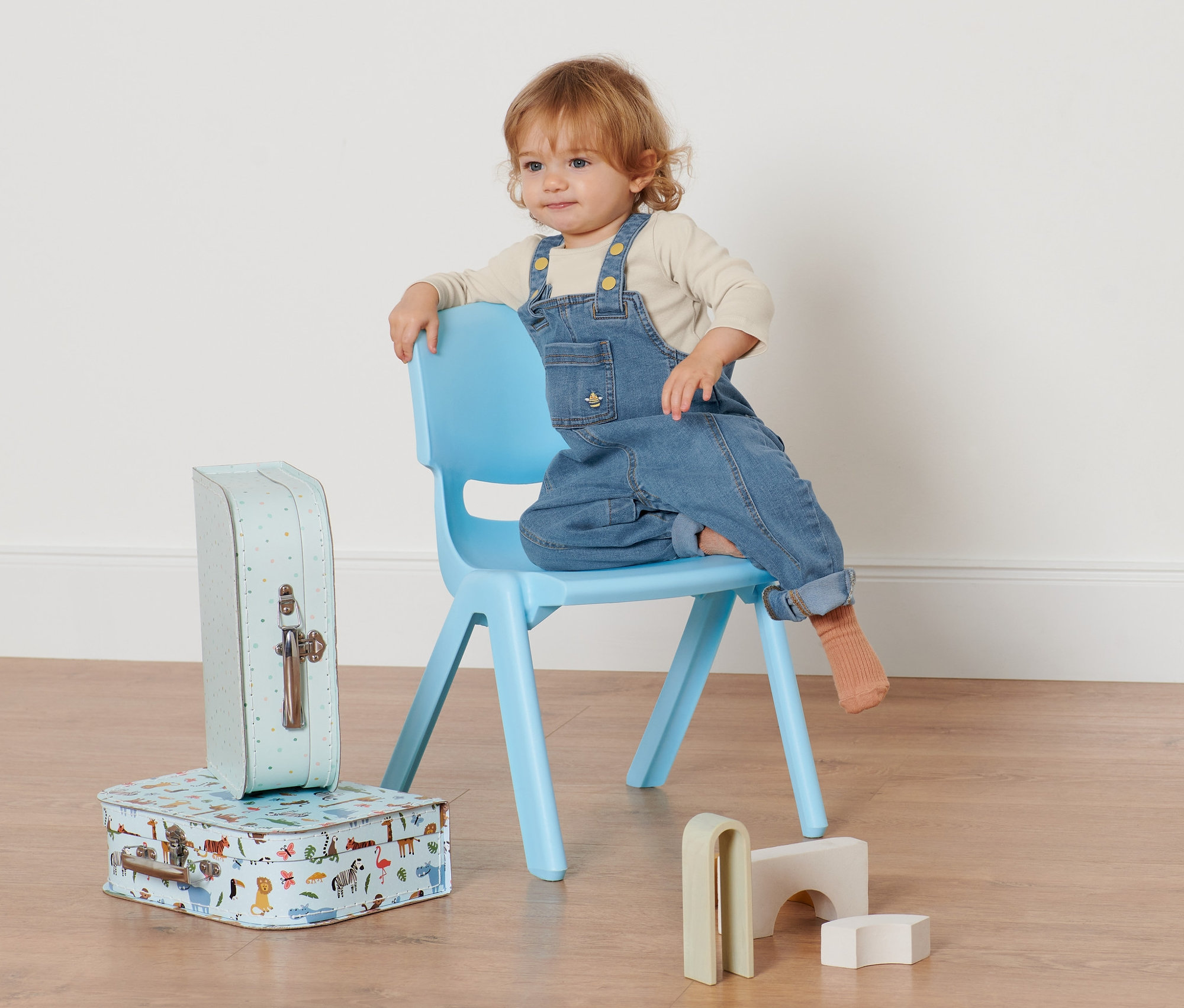 Batole sedí na modré židli a má na sobě džínové kalhoty s laclem. Vedle židle leží dvě kufříky a dřevěné hračky.