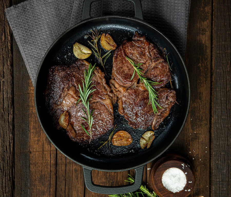 Dva steaky s rozmarýnem a česnekem v litinové servírovací pánvi »COMBEKK Sous-Chef« na dřevěném stole.