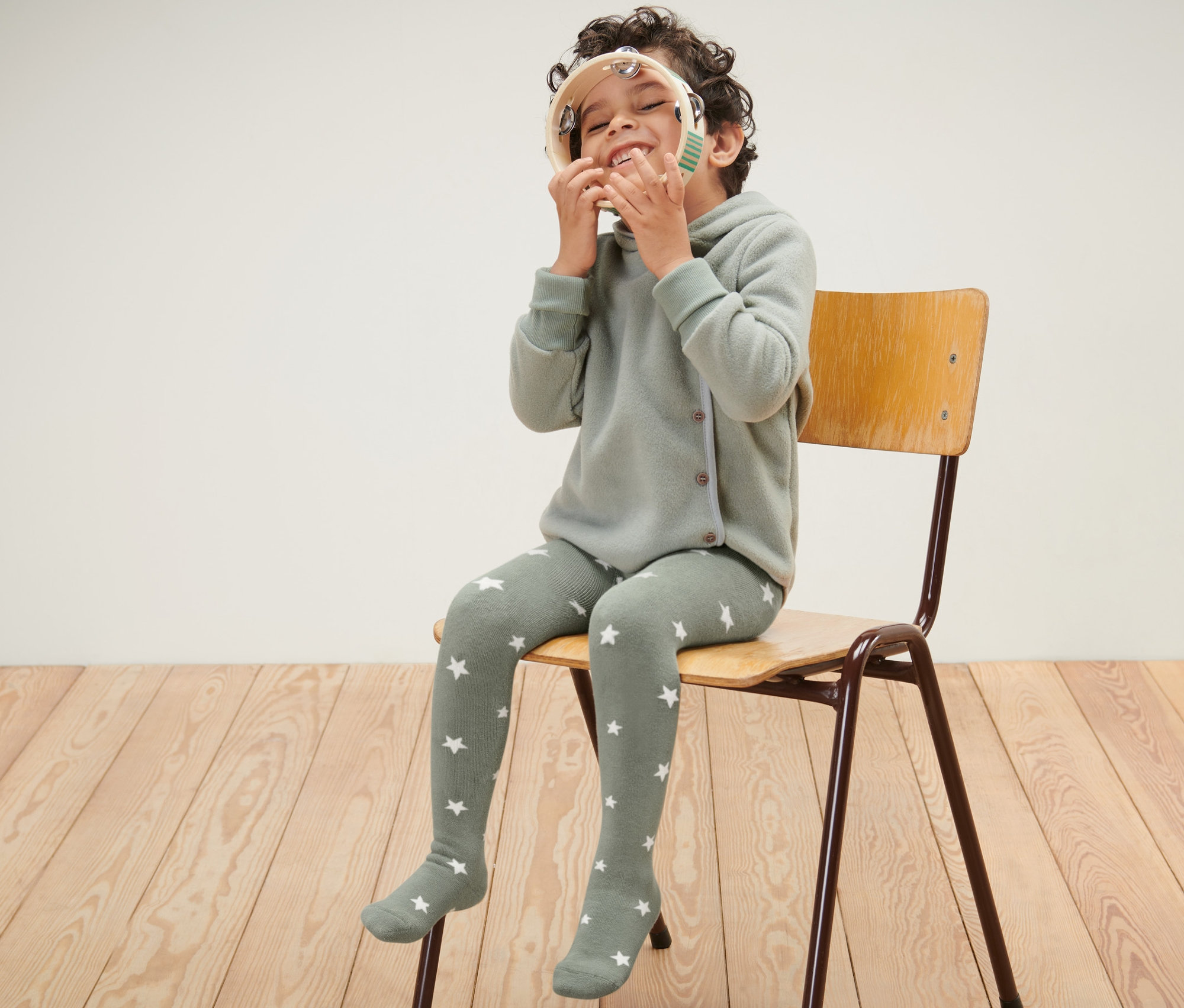 Chlapec sedí na židli a hraje si s tamburínou. Má na sobě modré a zelené dětské termo punčocháče.