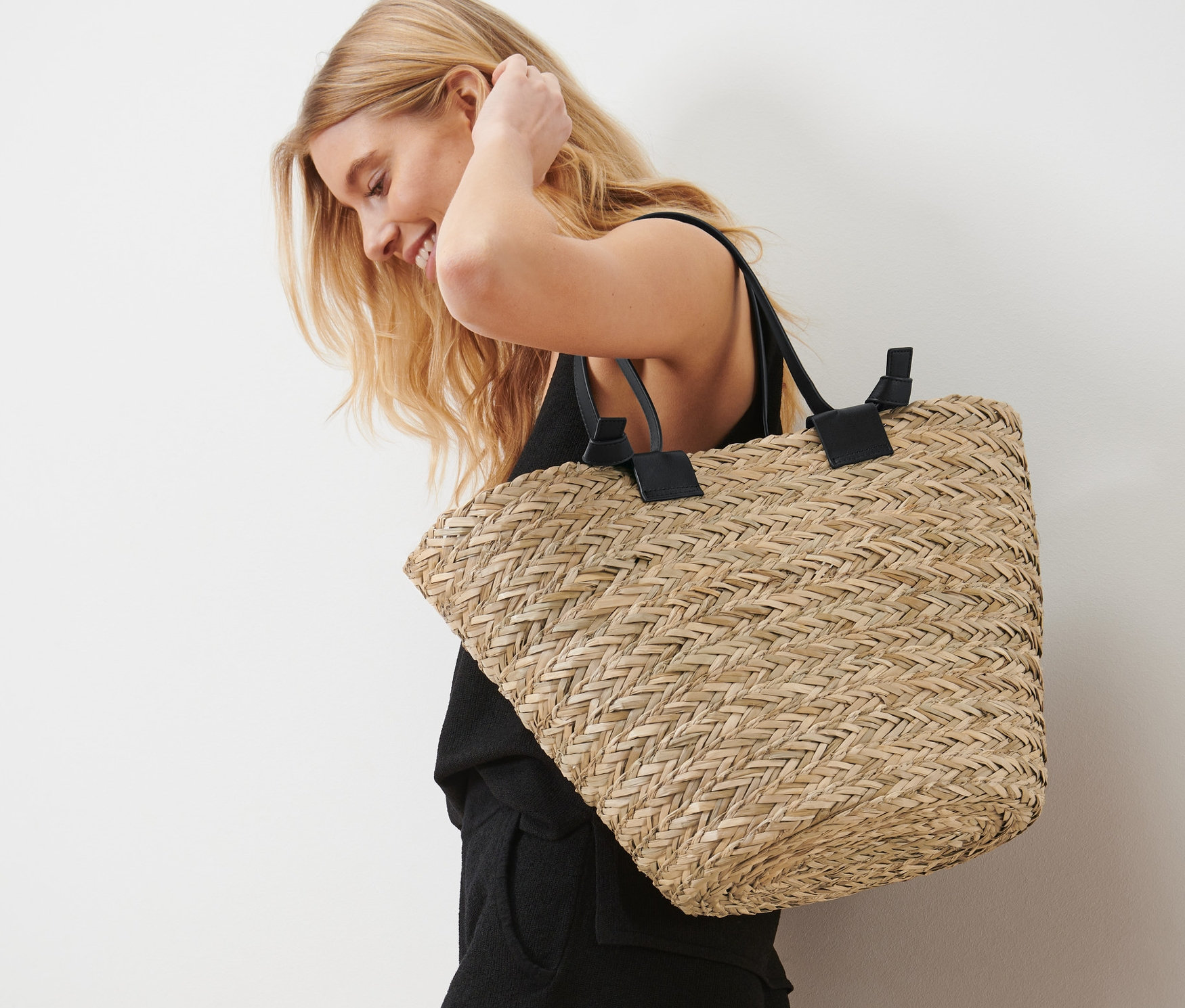 Blondýna se usmívá a má na rameni proutěnou tašku.