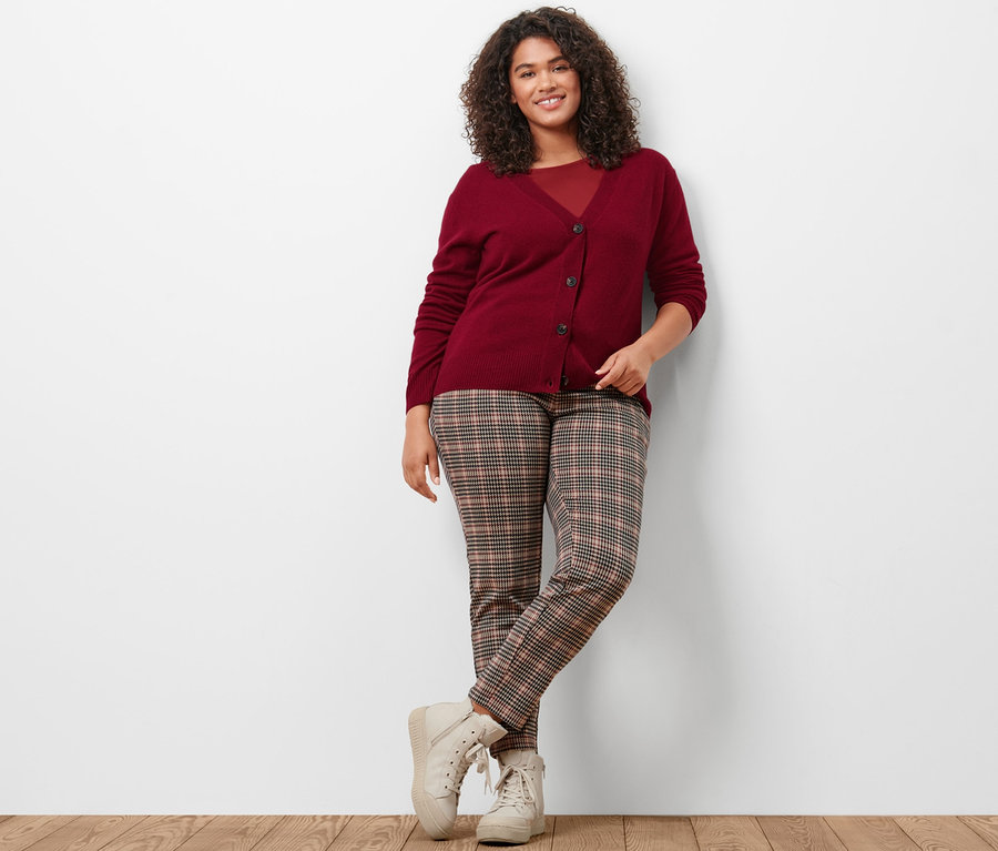 Žena pózuje v pleteném kabátku s kašmírem, vínově červeném triku s 3/4 rukávem a joggingových kalhotách se vzorem kohoutí stopy.