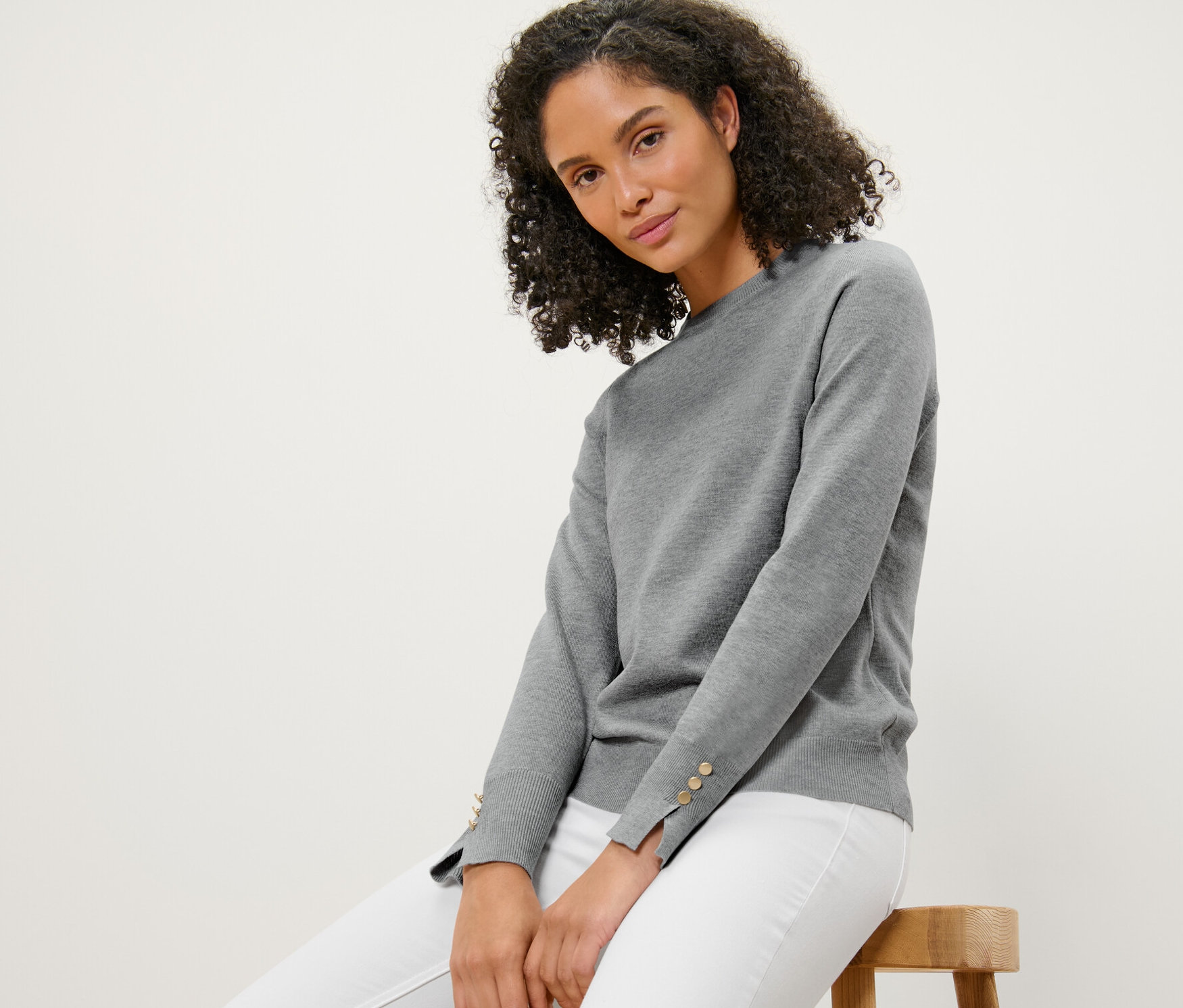 Žena sedí na stoličce a má na sobě šedý melírovaný svetr z jemného úpletu.