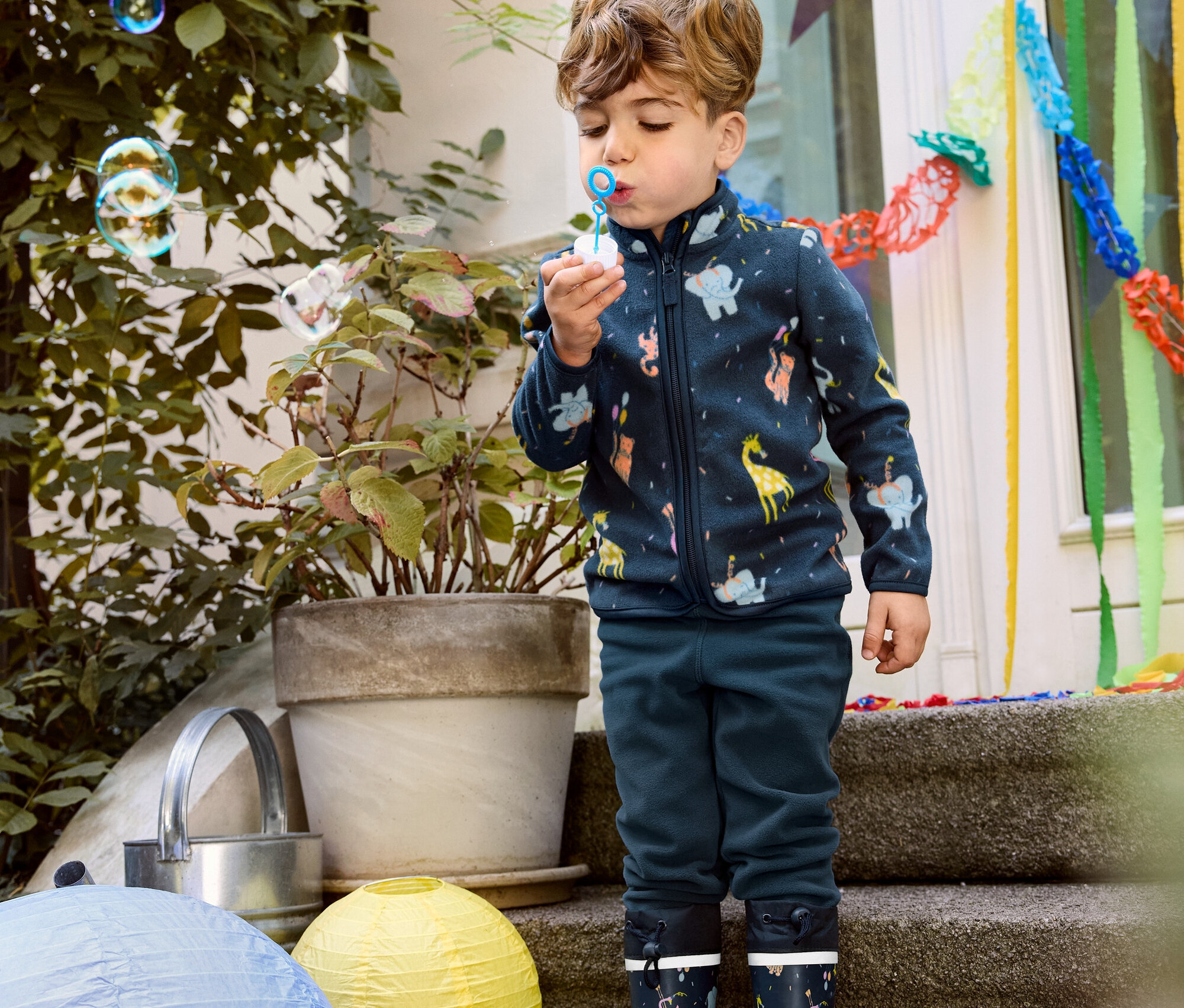 Chlapec v dětské fleecové bundě, fleecových kalhotách a gumových holínkách pouští bubliny.