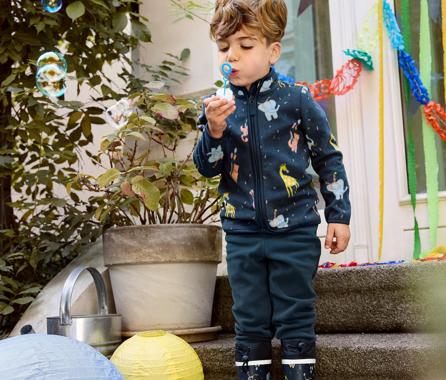 Chlapec v dětské fleecové bundě, fleecových kalhotách a gumových holínkách pouští bubliny.