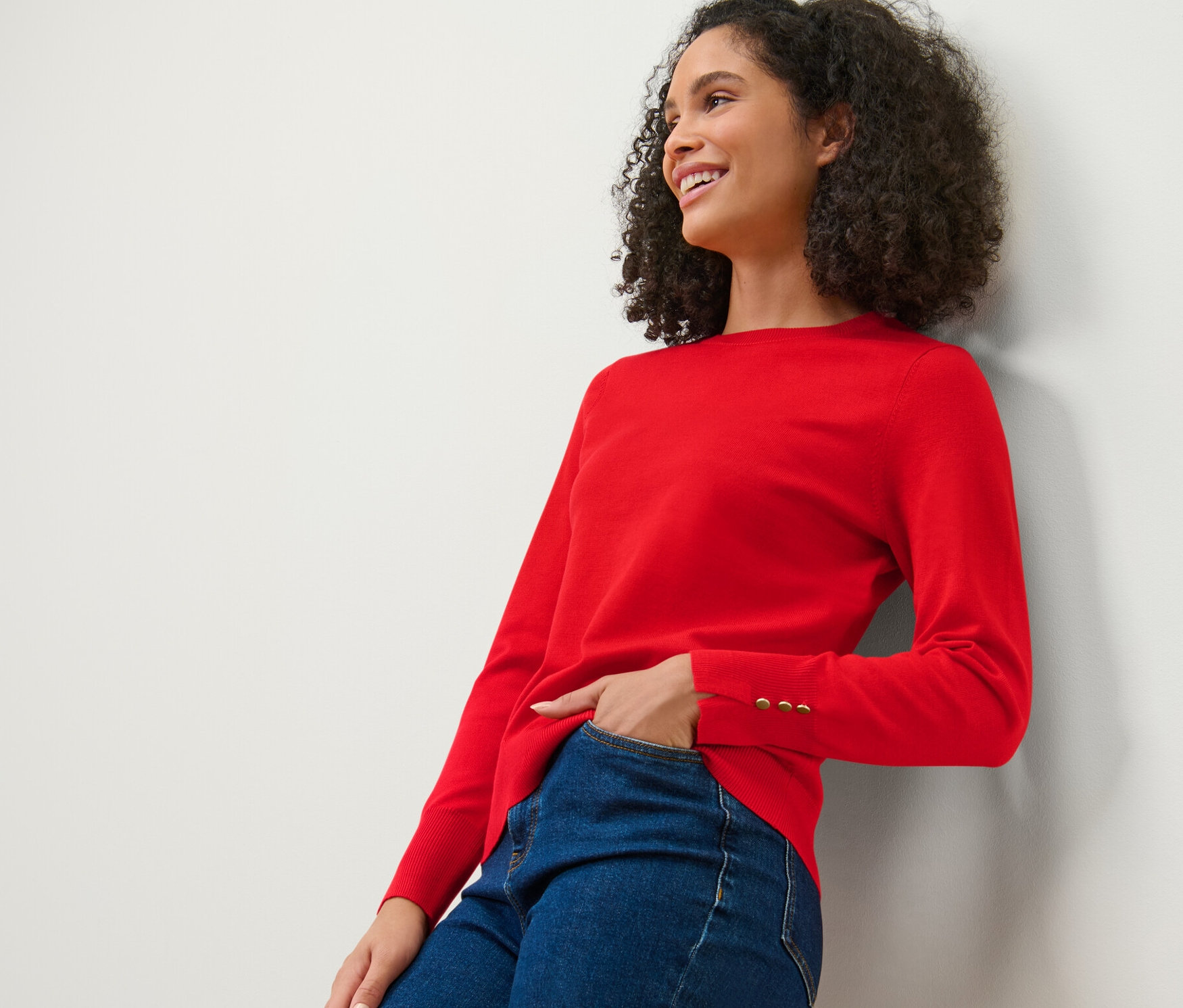 Usmívající se žena opřená o zeď má na sobě červený svetr a džíny.