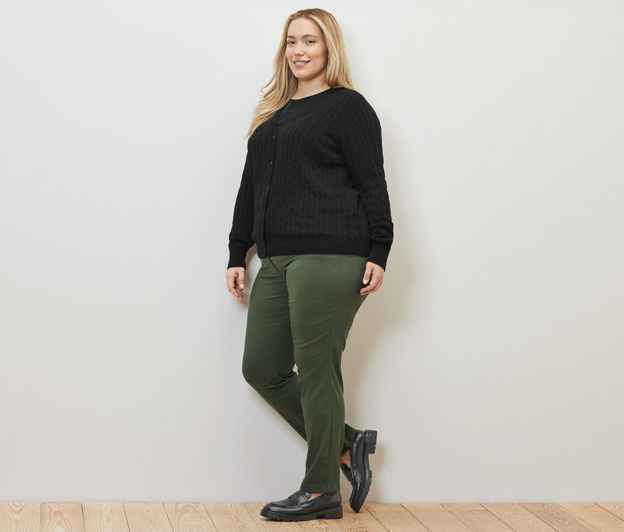 Žena pózuje v černém kardiganu z jemného úpletu s copánkovým vzorem a zelených bengalínových kalhotách.