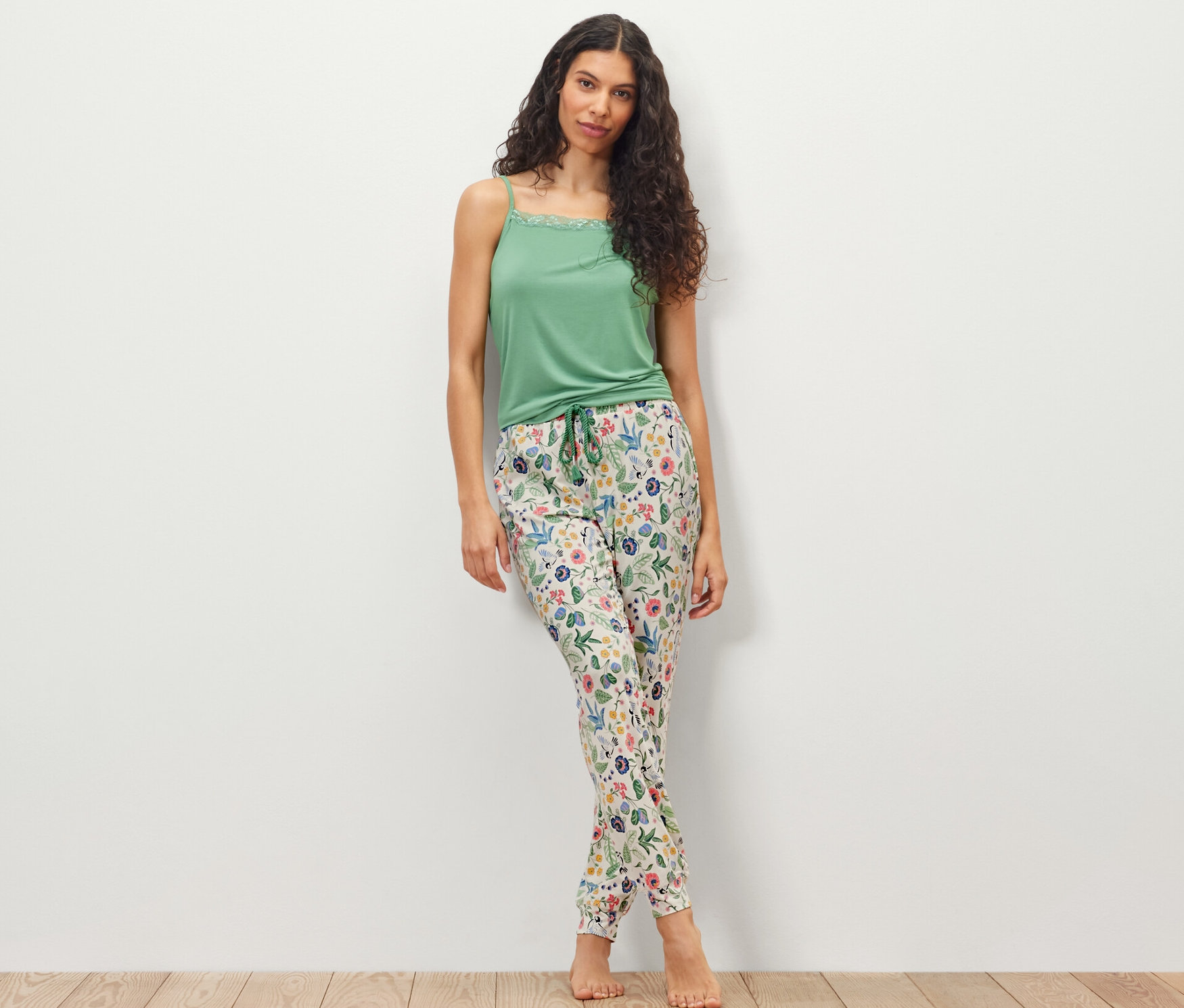 Žena stojící před bílou zdí má na sobě zelený top a pyžamové kalhoty.