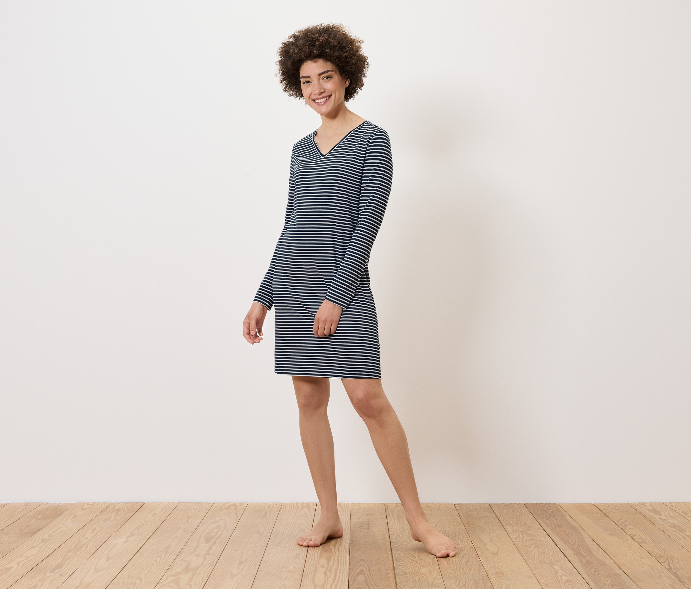 Žena stojí na dřevěné podlaze a má na sobě pruhované šaty s dlouhým rukávem.