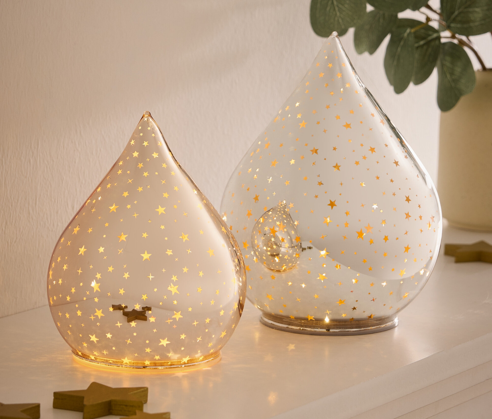 Dvě skleněné lampy ve tvaru slzy se zlatými hvězdami stojí na bílém povrchu vedle zelených dřevěných hvězd.