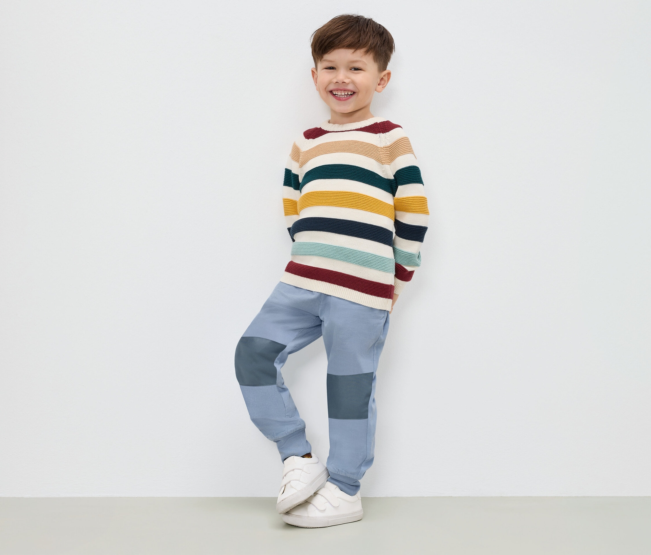 Chlapec v pruhovaném svetru, modrých kalhotách a bílých teniskách stojí u bílé stěny.