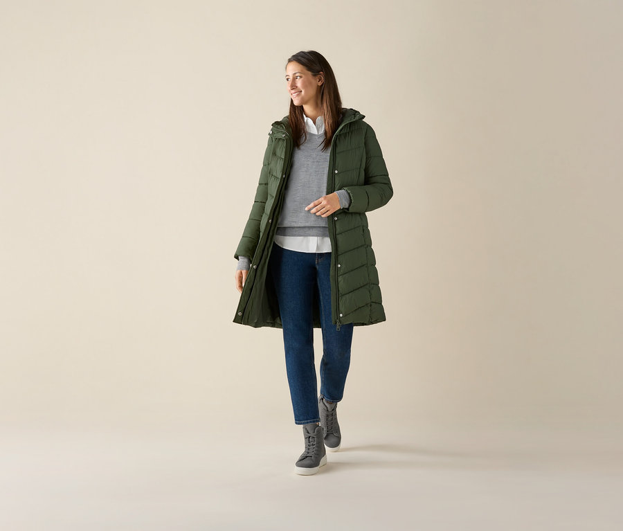 Žena má na sobě šedý svetr z jemného úpletu merino, zelenou bundu a džíny.