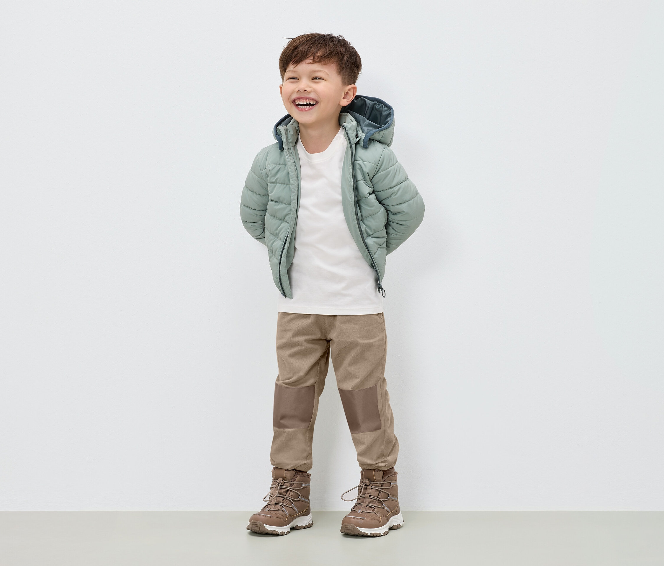 Usmívající se chlapec stojí v péřové bundě světle zelené barvy, béžových kalhotách a hnědých botách.