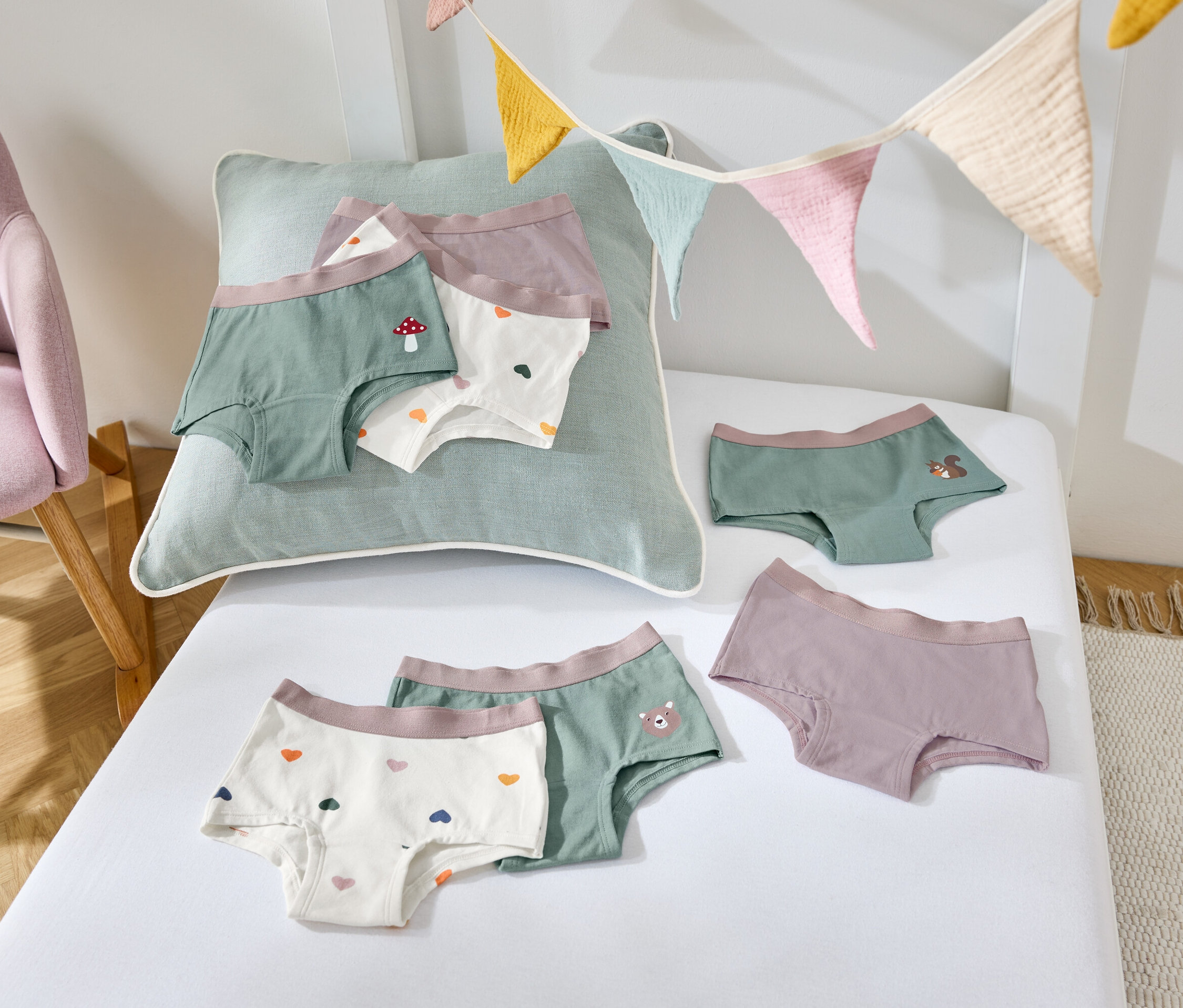 Na bílém prostěradle leží dětské kalhotky různých barev a vzorů, vedle stojí fialové křeslo.