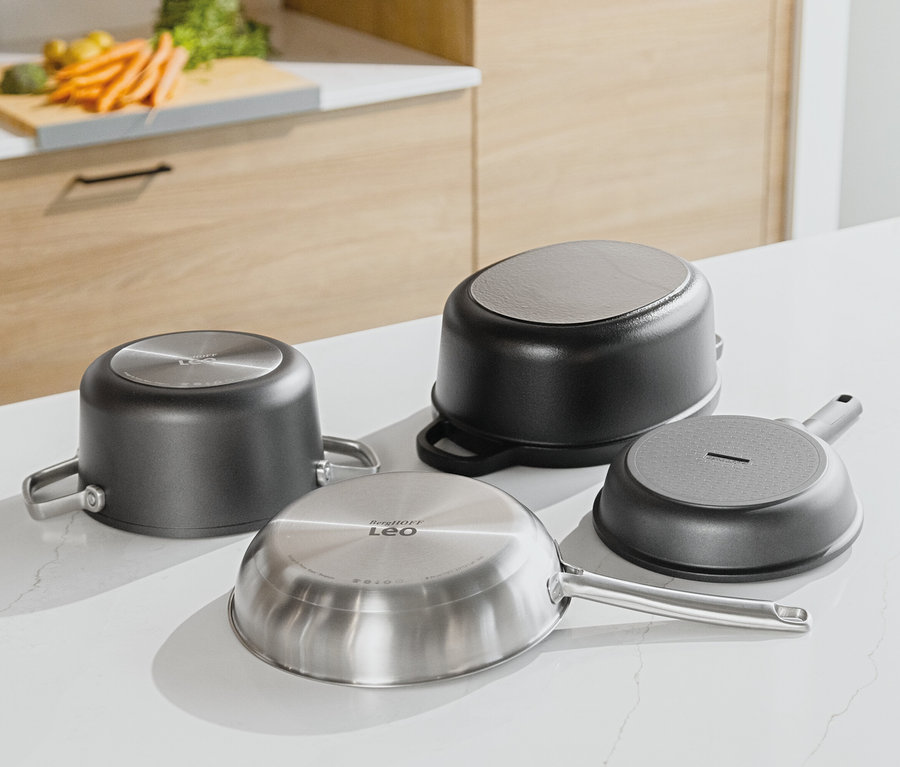 Čtyři pánve leží dnem vzhůru na bílé kuchyňské desce. V pozadí je vidět dřevěná kuchyňská linka a zelenina na prkénku.