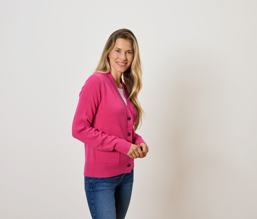 Žena s blond vlasy pózuje v růžovém svetru a modrých džínách.